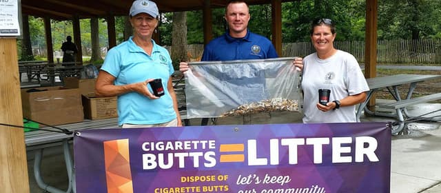Two women and a man stand behind a purple banner reading CIGARETTE BUTTS = LITTER, DISPOSE OF CIGARETTE BUTTS; the outer two hold small black pocket ashtrays and all three hold up a clear bag full of collected cigarette ends. A picnic shelter is behind them.