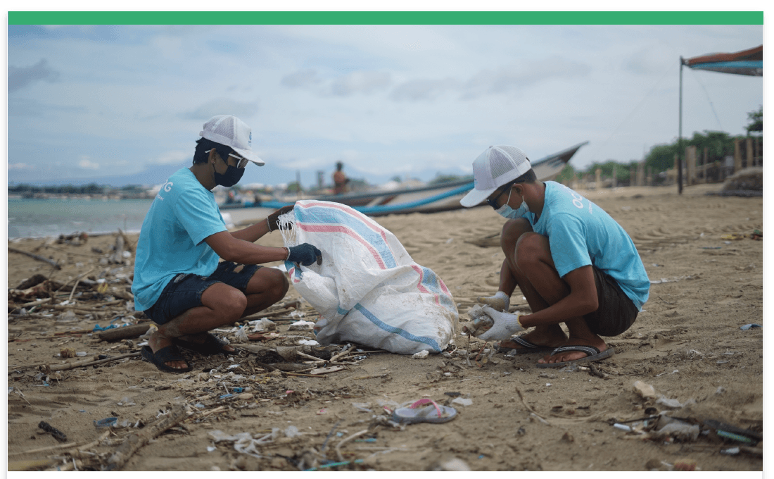 Two young men in pale blue t-shirts, caps, face masks and gloves crouch on a debris-strewn tropical beach, filling a large woven sack with litter; fishing boats and a canopy stand on the sand behind them.