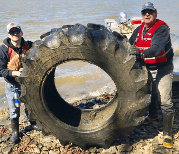 Two people in life jackets and waders stand on a rocky riverbank hauling an enormous muddy tractor tyre upright between them; a small boat with an outboard motor is pulled up behind on the brown water.