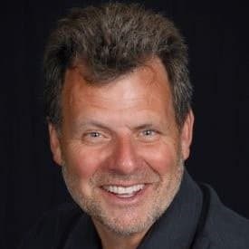 Square studio headshot of a smiling middle-aged man with short greying hair and a light beard, wearing a dark jacket over a lighter shirt against a black background.