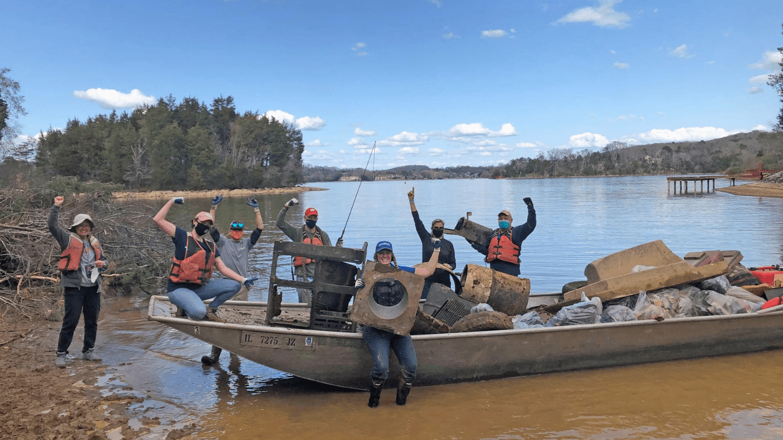 Seven volunteers in life jackets, gloves and face masks cheer with arms raised beside a flat-bottomed metal boat pulled up on a muddy lake shore, its hull loaded with a rusted drum, scrap metal, a foam seat and bags of collected rubbish; the boat is registered IL 7275 JZ.