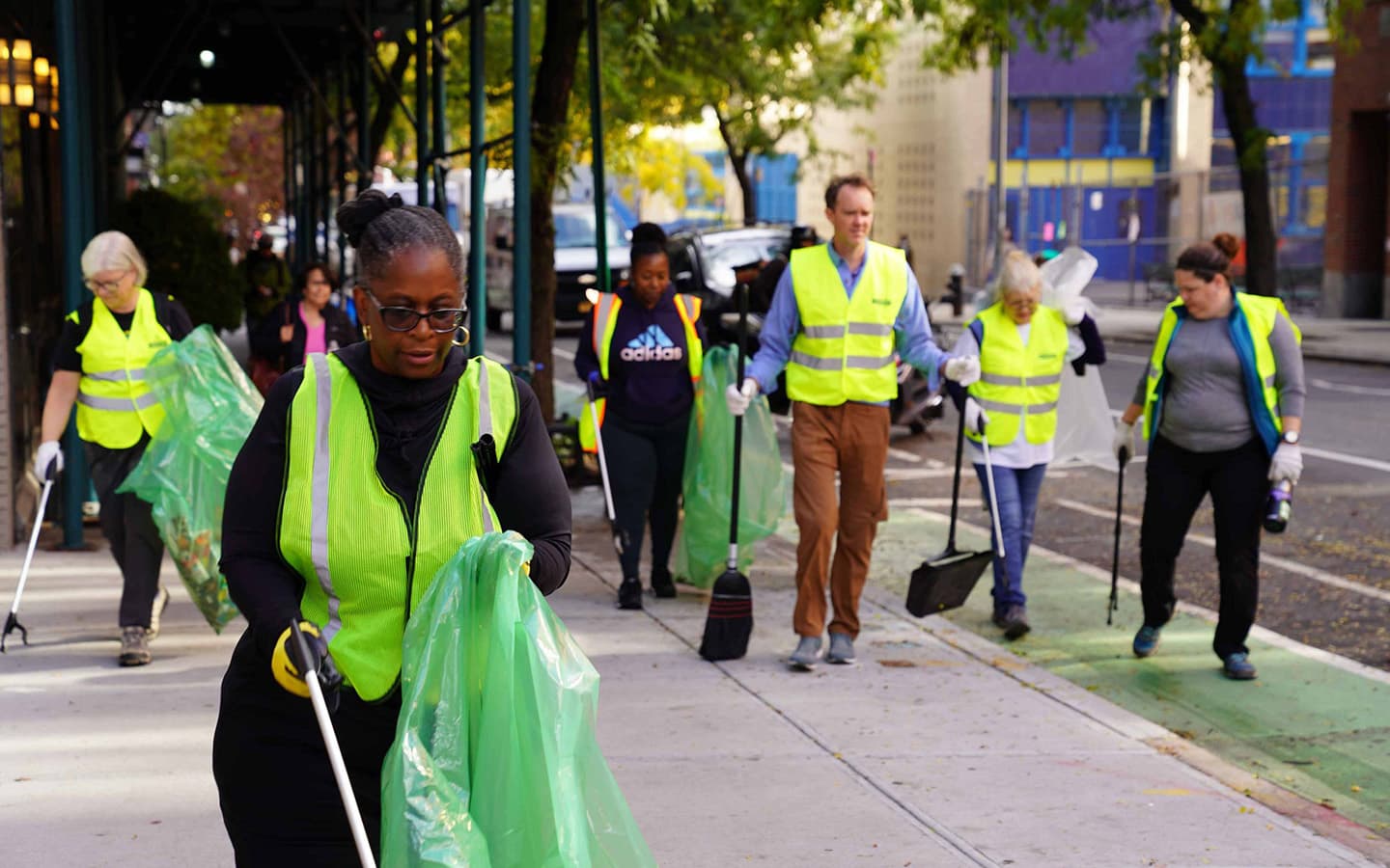 Cropped view of the same lime-vested clean-up line on a tree-lined city pavement, with the nearest volunteer in glasses and hoop earrings holding a green sack and grabber while others sweep and bag litter behind her.