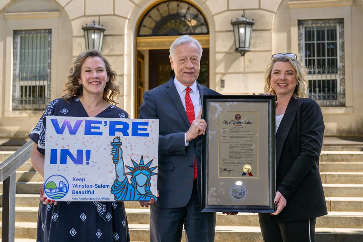 Three people stand on stone steps outside a columned civic building. One woman holds a board reading WE'RE IN! with a Keep Winston-Salem Beautiful logo and a Statue of Liberty drawing; a man in a suit and red tie and a woman in black hold a framed City of Winston-Salem proclamation with a gold seal.