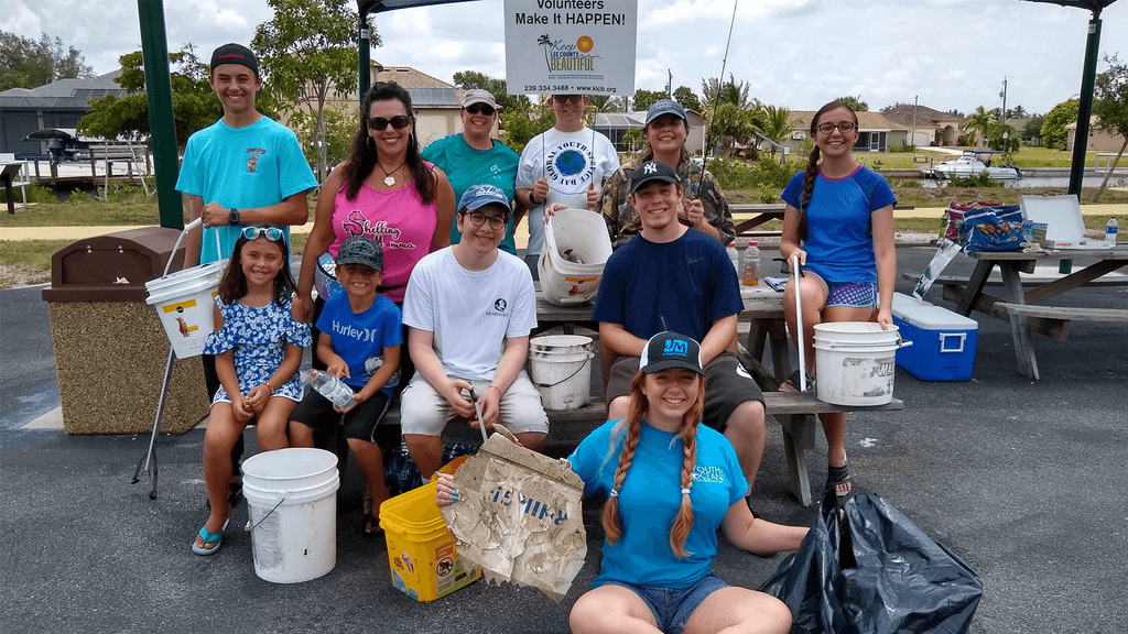 Twelve people of mixed ages, from small children to teenagers and adults, pose around a picnic table in a coastal car park with white buckets, a black sack and a large sheet of collected debris. A banner overhead reads Volunteers Make It HAPPEN! with a Keep Lee County Beautiful logo.