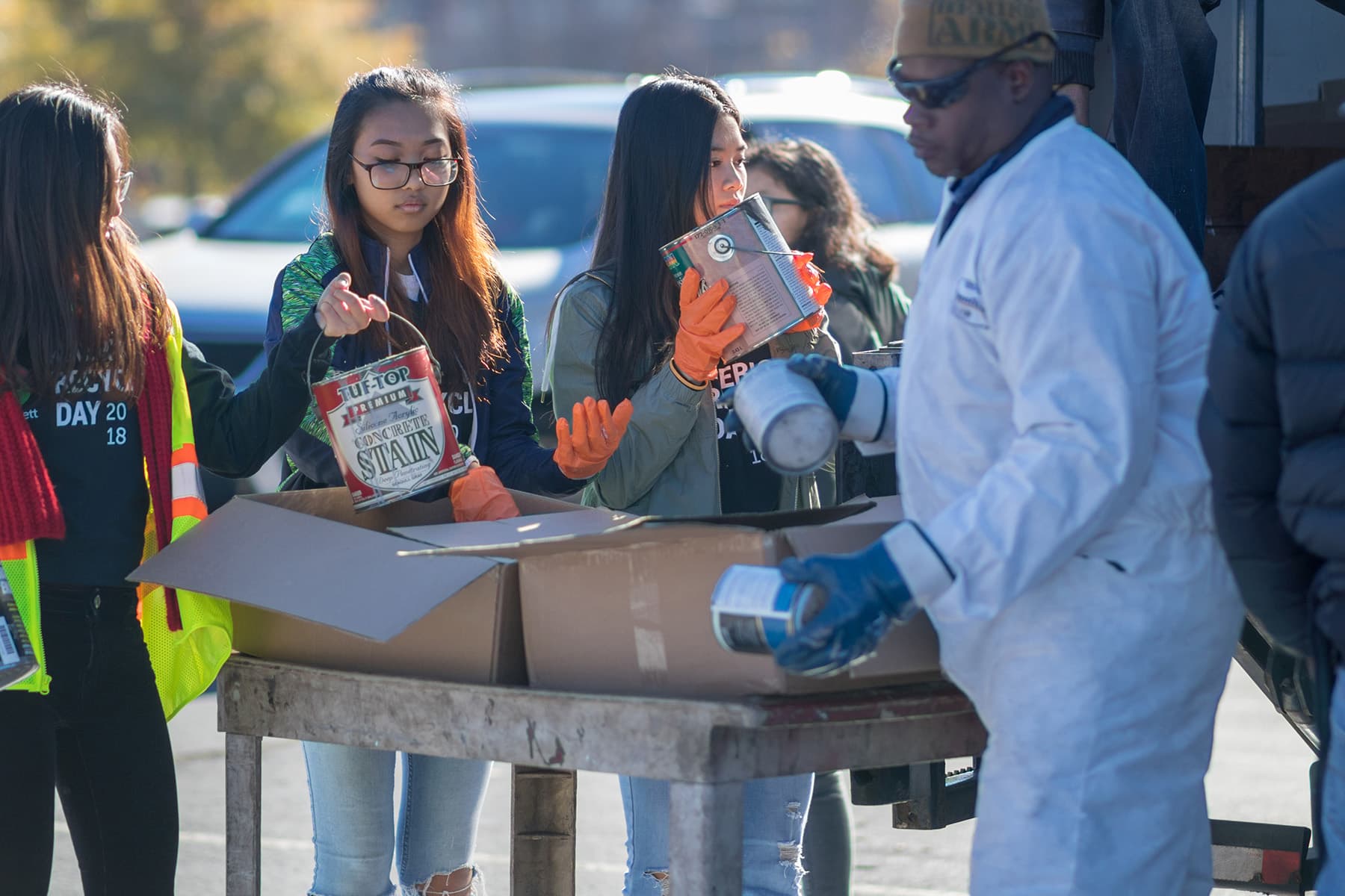 Volunteers at an outdoor America Recycles Day collection hand tins of concrete stain and paint across a trestle table into open cardboard boxes. One wears a hi-vis vest reading RECYCLE DAY 2018; a steward in white coveralls and blue gloves takes a can from them.