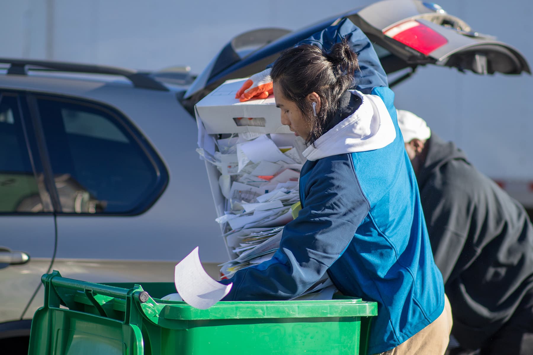 A volunteer in a blue jacket empties a boxful of shredded paper and envelopes into a green wheeled recycling cart at a drive-up collection point, with the open tailgate of a car behind her.