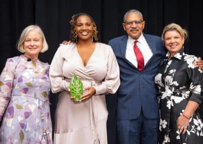 The City of Atlanta honoree, in a blush dress, holds a green glass leaf trophy on stage at the 2025 Power of Beauty celebration, beside a man in a dark suit and red tie.