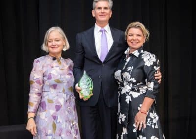 The Coca-Cola Company's honoree, at centre in a dark suit and lilac tie, holds a green glass leaf trophy on stage at the 2025 Power of Beauty celebration, flanked by two women in floral dresses.