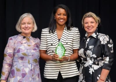 The Hilton honoree, at centre in a black-and-white striped blazer, holds a green glass leaf trophy on stage at the 2025 Power of Beauty celebration, flanked by two women in floral dresses.