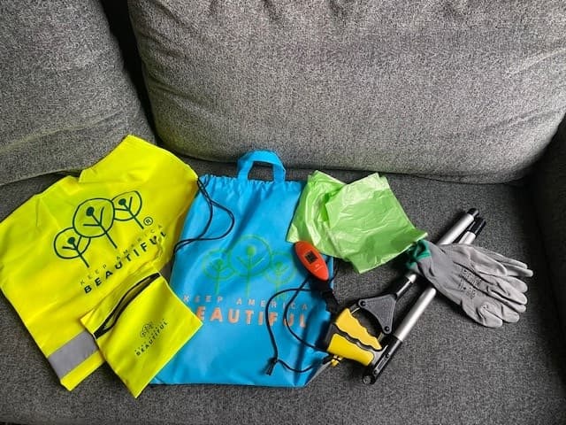 A Great American Cleanup volunteer kit laid out on a grey sofa: a lime hi-vis vest with a silver reflective stripe and the Keep America Beautiful tree logo, a blue Keep America Beautiful drawstring bag, a black and yellow long-handled litter grabber, a bundle of bright green liner bags, grey work gloves, and a small orange digital hanging scale.