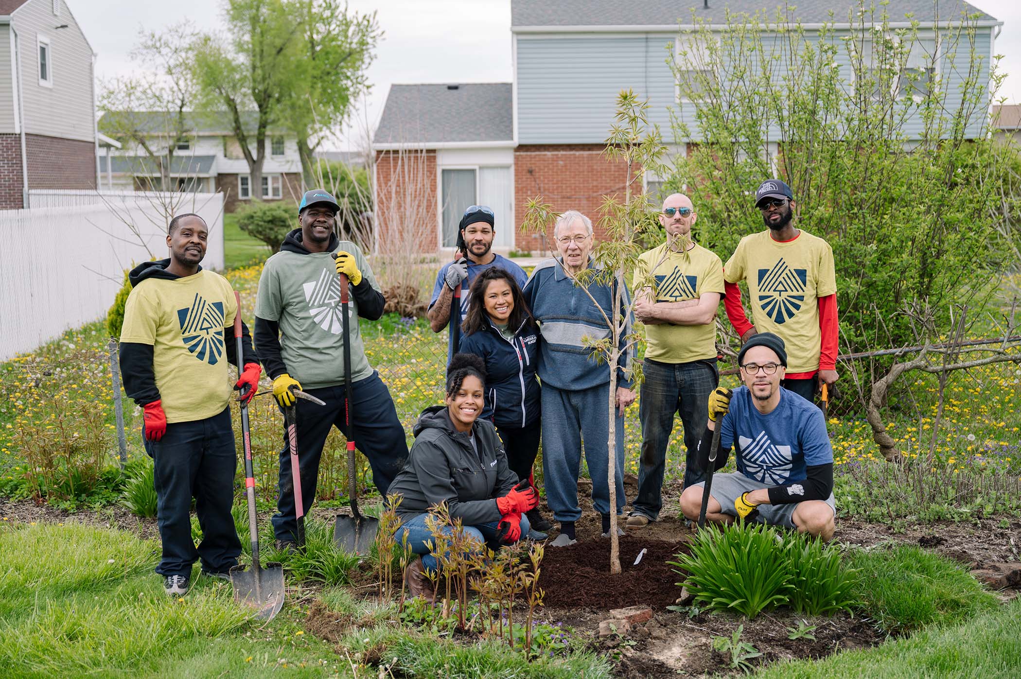 Nine volunteers, several in yellow and blue Team Rubicon t-shirts and holding shovels, gather smiling around a freshly mulched sapling in a suburban back garden, houses behind them.