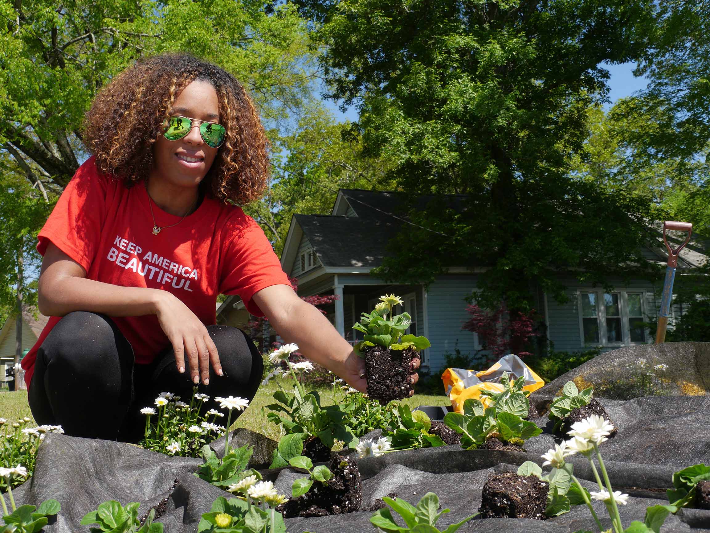 A volunteer in a red Keep America Beautiful t-shirt and sunglasses kneels to press a white-flowering plant into a landscaped bed outside a pale blue clapboard house.