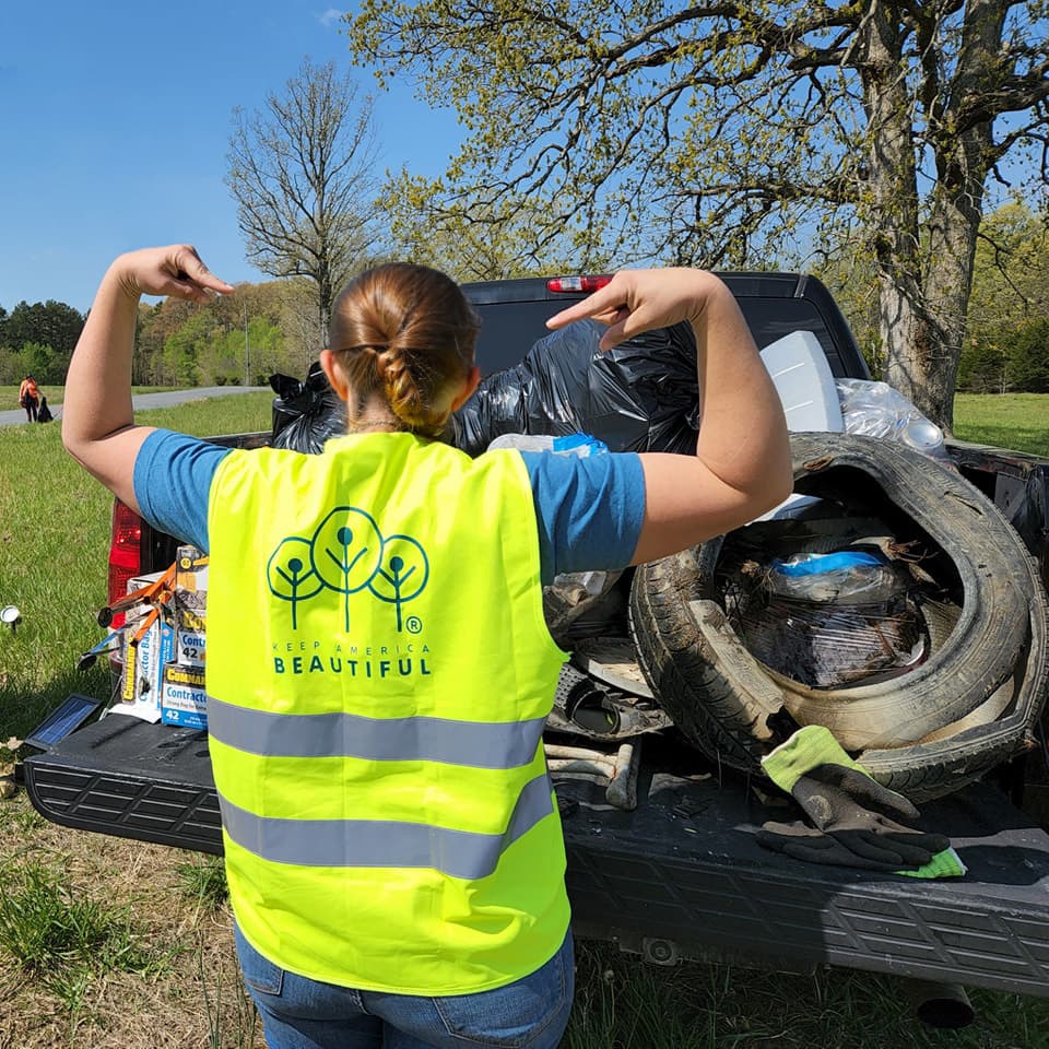 A volunteer in a neon-yellow Keep America Beautiful safety vest, the three-trees mark across her back, raises both arms toward a pickup bed loaded with bagged litter, a discarded tyre and scrap pulled from a rural roadside.