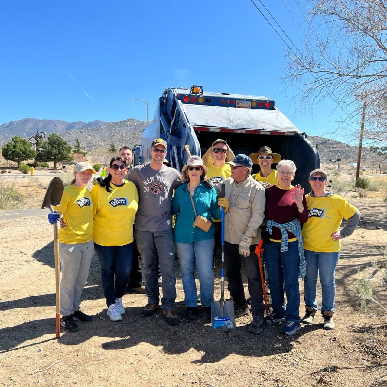 A large group of Keep Albuquerque Beautiful volunteers in yellow Cleanup Month shirts holds shovels and rakes in front of a city dump truck, mountains behind them under a clear desert sky.
