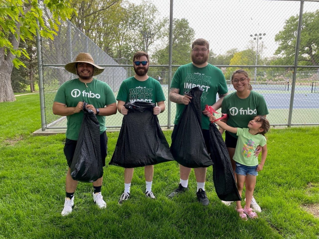 Four Keep Nebraska Beautiful volunteers in green FNBO t-shirts hold full black trash bags on a park lawn, a small child standing beside them.