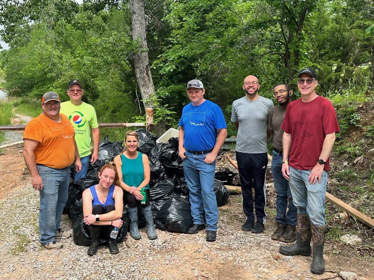 Eight Keep Oklahoma Beautiful volunteers stand beside a tall pile of filled black trash bags on a gravel path at a wooded lakeside.