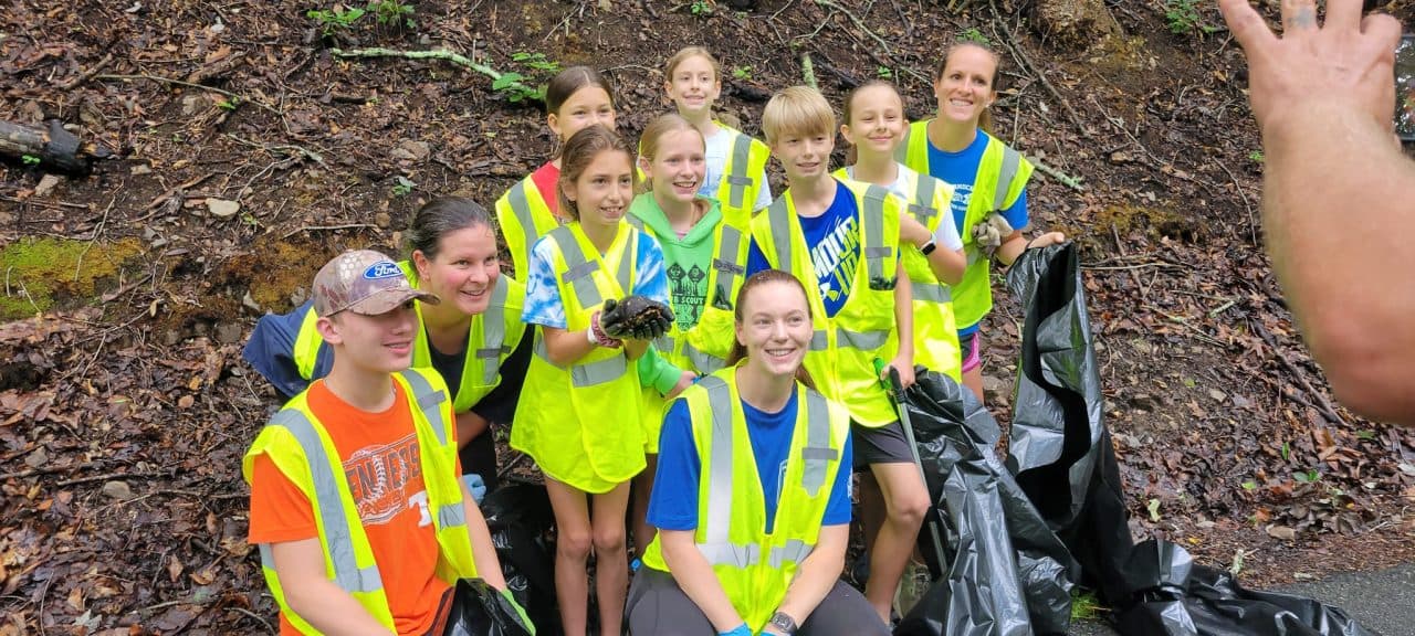 Kids and adults in neon-yellow safety vests kneel together on a wooded slope during a Keep Rome-Floyd Beautiful cross-country cleanup, holding work gloves and trash bags.